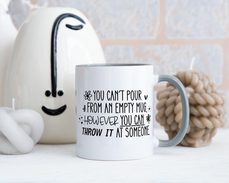 May include: White ceramic mug with a gray handle and rim, featuring the text "YOU CAN'T POUR FROM AN EMPTY MUG... HOWEVER YOU CAN THROW IT AT SOMEONE" in black. The mug is surrounded by decorative candles and a ceramic face.