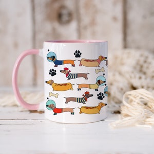 May include: White ceramic mug with a pink handle and rim. The mug features a colorful pattern of dachshunds wearing different outfits, including a space suit, a sailor hat, and a beret. The dachshunds are holding hot dogs and bones.