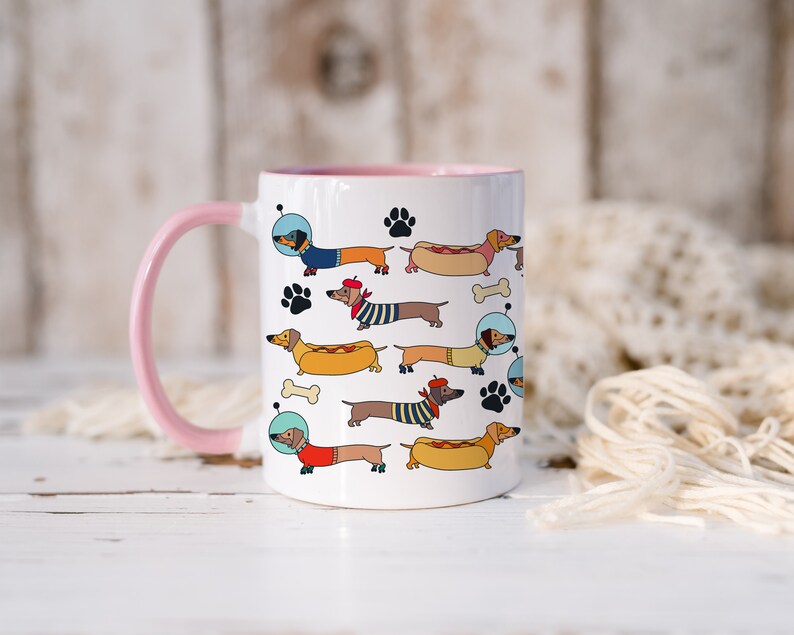 May include: White ceramic mug with a pink handle and rim. The mug features a colorful pattern of dachshunds wearing different outfits, including a space suit, a sailor hat, and a beret. The dachshunds are holding hot dogs and bones.