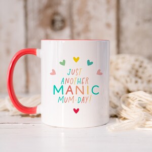 May include: A white ceramic mug with a bright red handle and interior. The front features the text "JUST ANOTHER MANIC MUM-DAY!" in colorful letters, surrounded by small, multi-colored heart icons arranged in a semi-circle above and below the text.