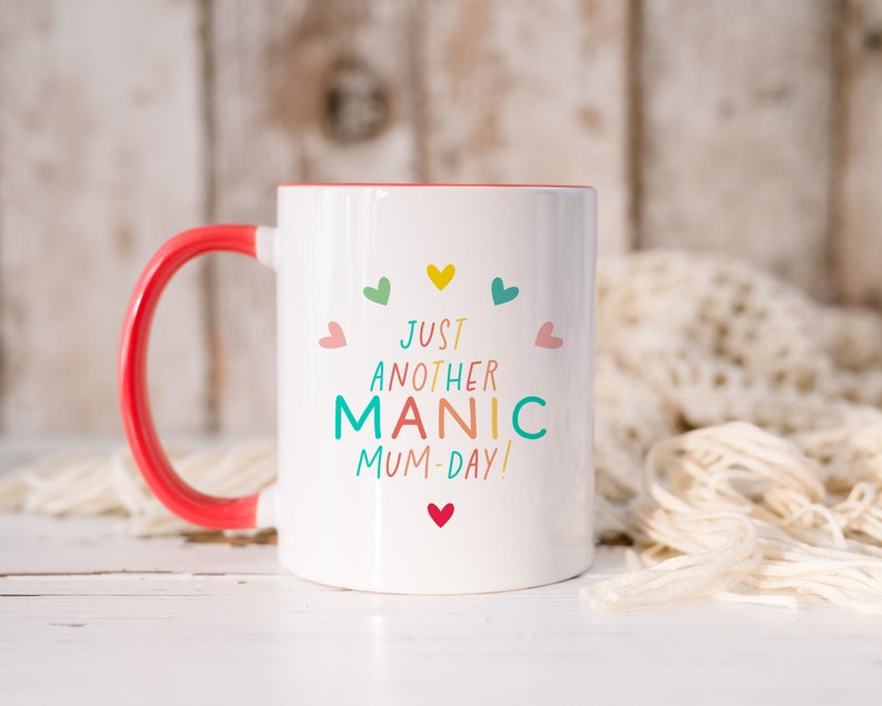 May include: A white ceramic mug with a bright red handle and interior. The front features the text "JUST ANOTHER MANIC MUM-DAY!" in colorful letters, surrounded by small, multi-colored heart icons arranged in a semi-circle above and below the text.