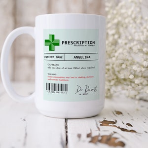 May include: White ceramic mug with a green cross and the text "PRESCRIPTION" and "ANGELINA". The mug has a black barcode and the text "CAFFEINE" and "take one dose of at least 250ml when required".