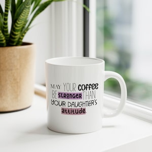 Peut inclure: Une tasse à café en céramique blanche avec le texte "May your coffee be stronger than your daughter's attitude." en lettres noires et violettes.