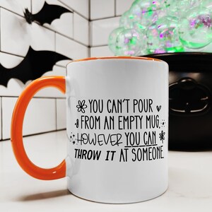May include: White ceramic mug with an orange handle and interior. The mug features black text that reads "YOU CAN'T POUR FROM AN EMPTY MUG, HOWEVER YOU CAN THROW IT AT SOMEONE." The mug is set against a Halloween-themed background.