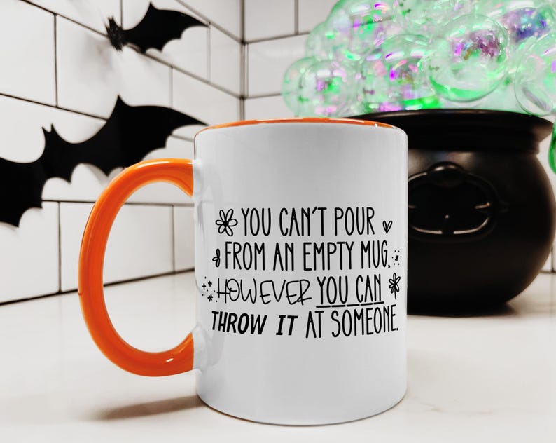 May include: White ceramic mug with an orange handle and interior. The mug features black text that reads "YOU CAN'T POUR FROM AN EMPTY MUG, HOWEVER YOU CAN THROW IT AT SOMEONE." The mug is set against a Halloween-themed background.