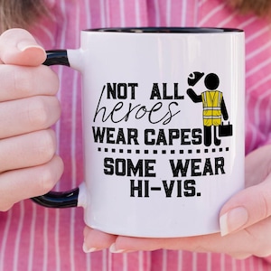Op de afbeelding: Een witte keramische mok met een zwarte handgreep en binnenrand. De mok is voorzien van zwarte tekst "NOT ALL heroes WEAR CAPES SOME WEAR HI-VIS." en een afbeelding van een stokfiguur in een geel reflecterend veiligheidsvest en een helm.