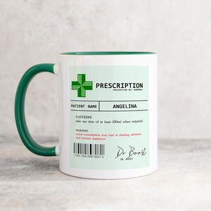 May include: A white ceramic mug with a green rim and handle. The mug has a green prescription label with the text "PRESCRIPTION" and "ANGELINA" printed on it. The label also includes a warning about excess consumption of caffeine.