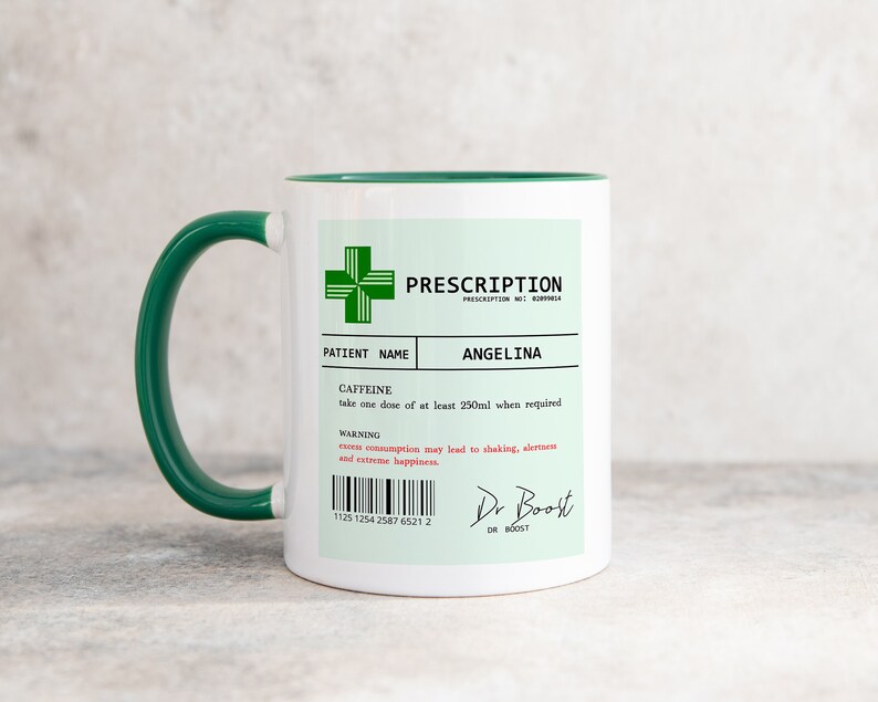May include: A white ceramic mug with a green rim and handle. The mug has a green prescription label with the text "PRESCRIPTION" and "ANGELINA" printed on it. The label also includes a warning about excess consumption of caffeine.