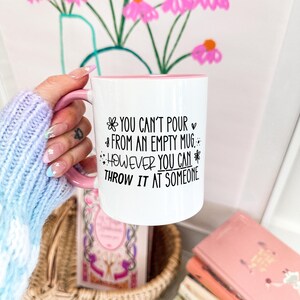 May include: White ceramic mug with a pink handle and interior. The mug features the text "YOU CAN'T POUR FROM AN EMPTY MUG. HOWEVER YOU CAN THROW IT AT SOMEONE" in black script. The mug is held in front of a floral painting.