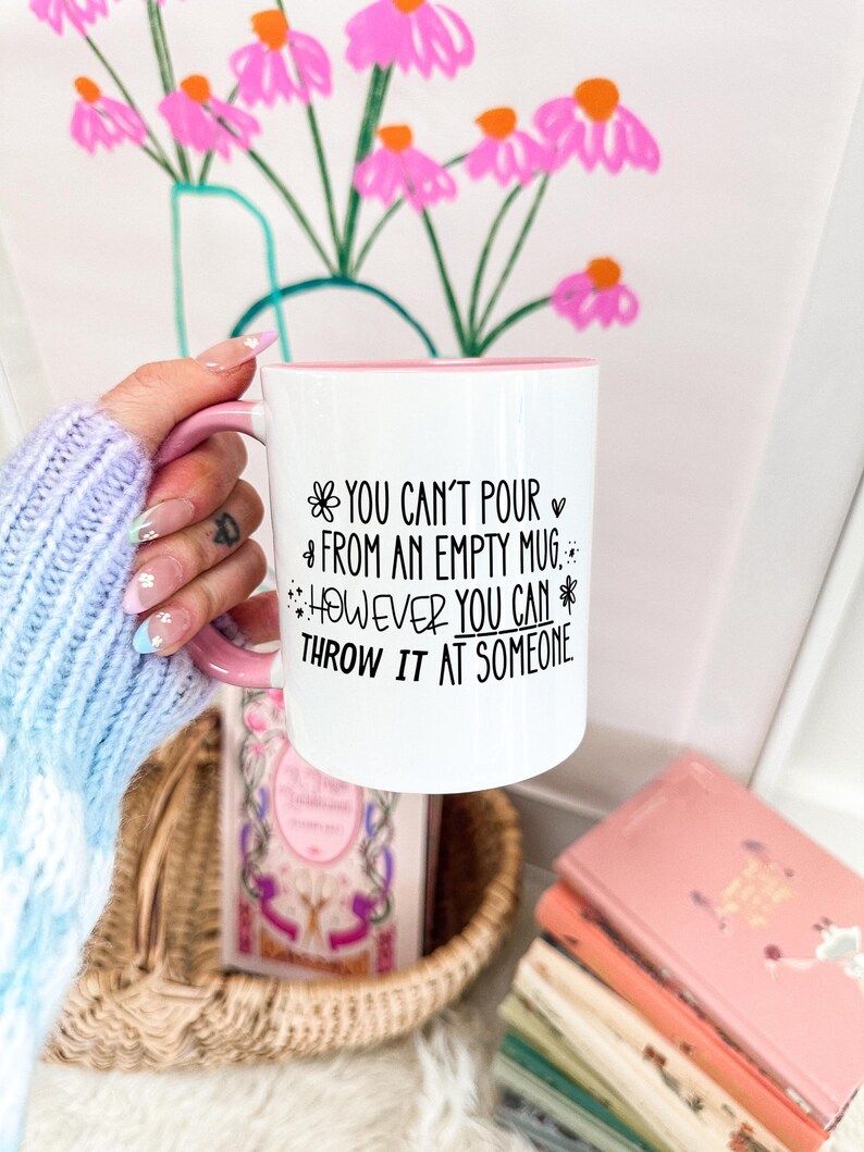 May include: White ceramic mug with a pink handle and interior. The mug features the text "YOU CAN'T POUR FROM AN EMPTY MUG. HOWEVER YOU CAN THROW IT AT SOMEONE" in black script. The mug is held in front of a floral painting.