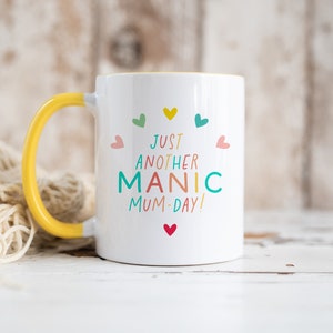 May include: A white ceramic mug with a bright yellow handle and interior. The front features the colorful text "JUST ANOTHER MANIC MUM-DAY!" arranged in a semi-circle, surrounded by small, multi-colored heart icons in pink, teal, yellow, and red.