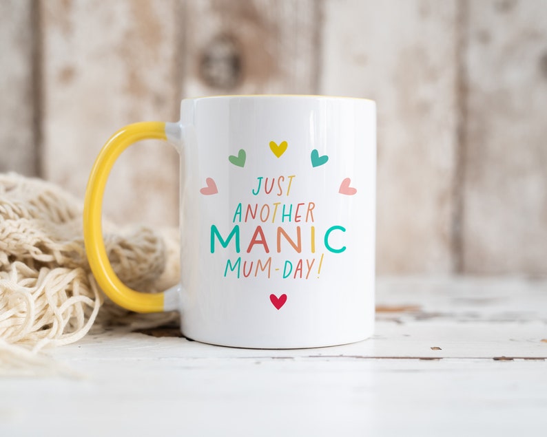 May include: A white ceramic mug with a bright yellow handle and interior. The front features the colorful text "JUST ANOTHER MANIC MUM-DAY!" arranged in a semi-circle, surrounded by small, multi-colored heart icons in pink, teal, yellow, and red.
