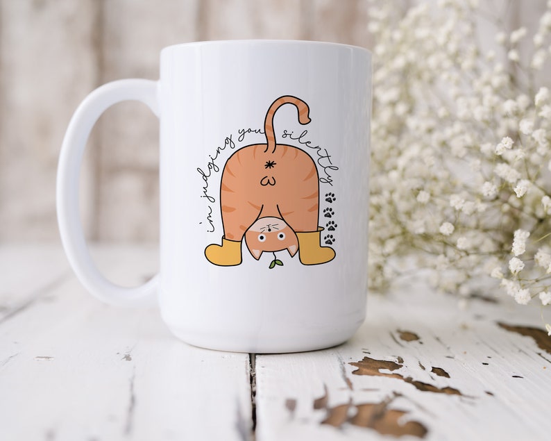 May include: White ceramic coffee mug with a cartoon orange cat wearing yellow boots and the text "I'm judging you silently" in black cursive.