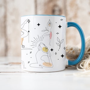 May include: White ceramic mug with a blue handle and rim. The mug features black line art illustrations of yoga poses, with yellow and pink accents. The design also includes decorative stars and leaves.