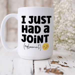 May include: A white ceramic mug with a handle featuring the black text I JUST HAD A JOINT (replacement) and a small yellow winking smiley face icon with pink cheeks printed on the front surface of the mug in a clear and legible font style.