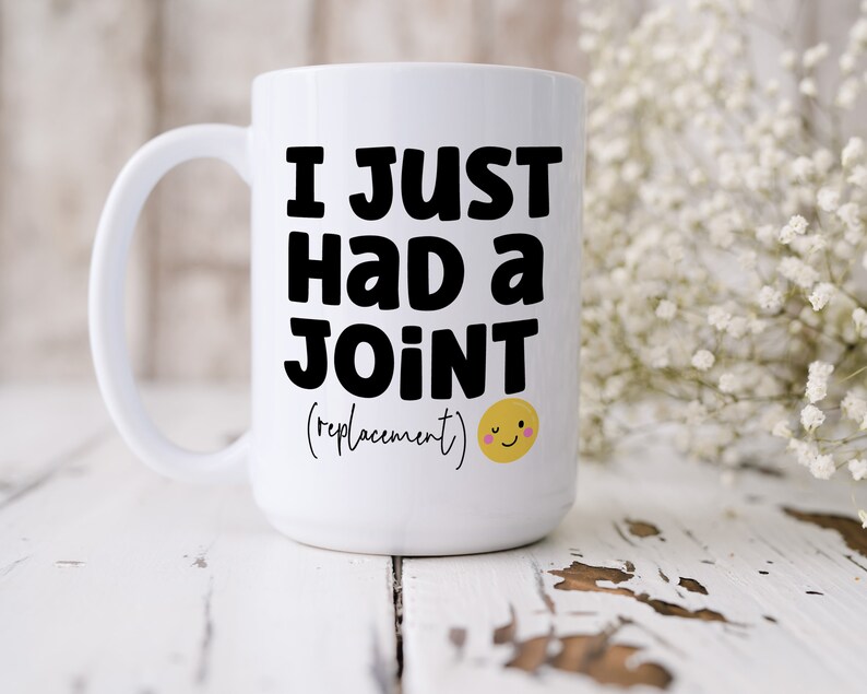 May include: A white ceramic mug with a handle featuring the black text I JUST HAD A JOINT (replacement) and a small yellow winking smiley face icon with pink cheeks printed on the front surface of the mug in a clear and legible font style.