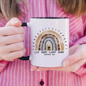 May include: White ceramic mug with a black handle. The mug features a watercolour rainbow with a cross in the centre and the text "Let your light shine Matthew 5:16".