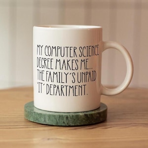 May include: A white ceramic mug with the text "MY COMPUTER SCIENCE DEGREE MAKES ME... THE FAMILY'S UNPAID 'IT' DEPARTMENT." The mug sits on a round, green marble coaster. The mug has a curved handle.