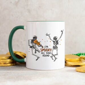 May include: White ceramic mug with a green rim and handle. The mug features a black and white illustration of two skeletons dancing. One skeleton is playing a violin. The text "I'm Spooky All Year Round" is printed on the mug.
