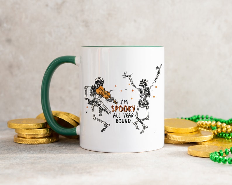 May include: White ceramic mug with a green rim and handle. The mug features a black and white illustration of two skeletons dancing. One skeleton is playing a violin. The text "I'm Spooky All Year Round" is printed on the mug.
