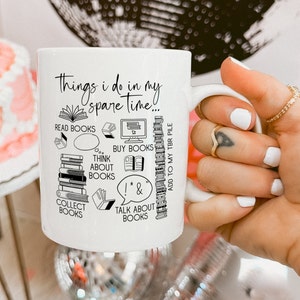 Op de afbeelding: Een witte keramische mok met een zwart ontwerp dat "Things I do in my spare time..." zegt en activiteiten gerelateerd aan het lezen van boeken opsomt. De activiteiten omvatten "Read Books", "Collect Books", "Buy Books", "Think About Books", "Talk About Books" en "Add to my TBR pile".