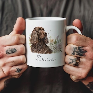 May include: White ceramic mug with a watercolour illustration of a brown dog with long, wavy fur, and floral accents. The name "Eric" is written in cursive below the dog. The mug has a standard handle.