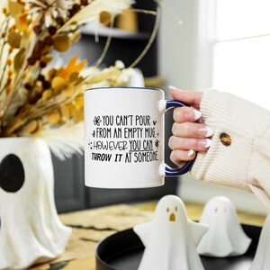 May include: White ceramic mug with a blue rim and handle, featuring the text "You can't pour from an empty mug, however you can throw it at someone." The mug is held in front of a decorative vase and ghost figurines.