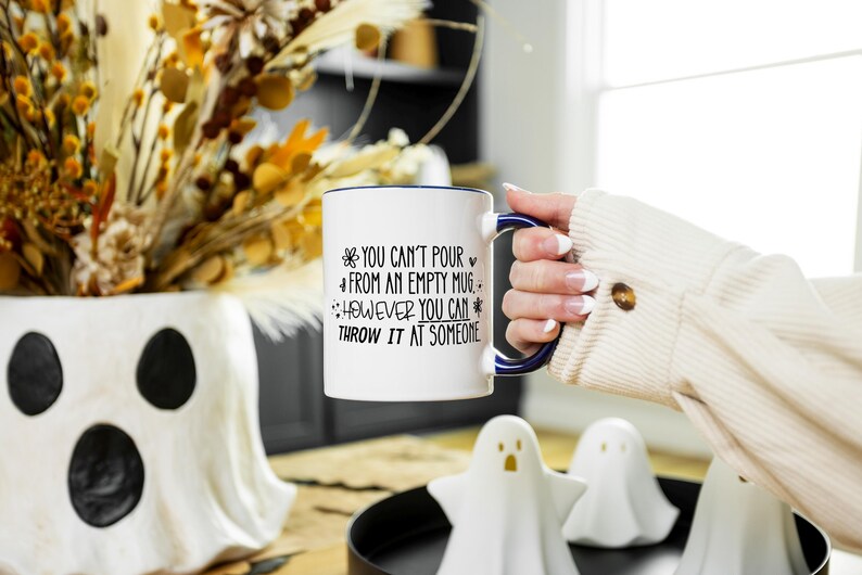 May include: White ceramic mug with a blue rim and handle, featuring the text "You can't pour from an empty mug, however you can throw it at someone." The mug is held in front of a decorative vase and ghost figurines.