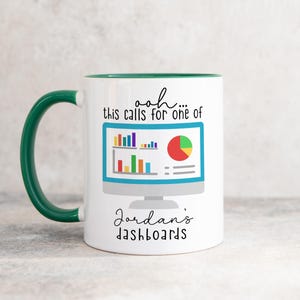 May include: A white ceramic mug with a dark green handle and interior. The front features a graphic of a computer monitor displaying colorful bar and pie charts, with the text "ooh... this calls for one of Jordan's dashboards" in black script.