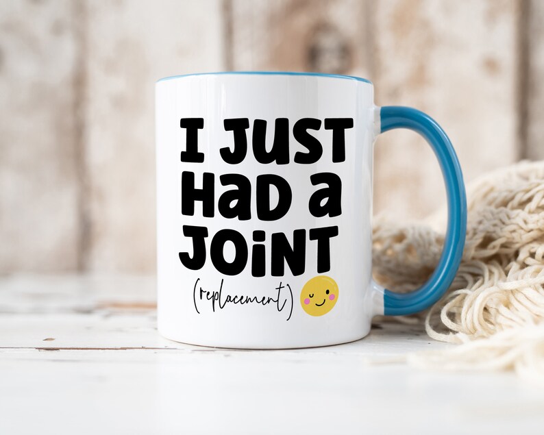 May include: A white ceramic mug with a blue interior and handle. The front features bold black text that reads I JUST HAD A JOINT (replacement) followed by a small yellow winking smiley face icon with pink cheeks on the lower right side.