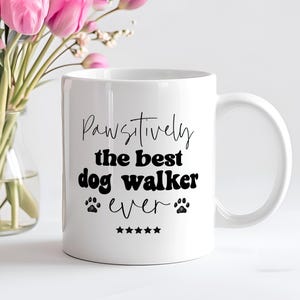 Può includere: Tazza da caffè in ceramica bianca con testo nero che recita "Pawsitively the best dog walker ever" con un'impronta di zampa su entrambi i lati della parola "ever" e cinque stelle sotto.