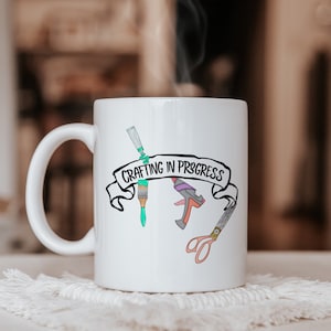 May include: A white ceramic mug featuring a black banner graphic with the text "CRAFTING IN PROGRESS" surrounded by illustrations of a paint tube, a paintbrush, a glue gun, and a pair of scissors in various colours including teal, orange, and grey.