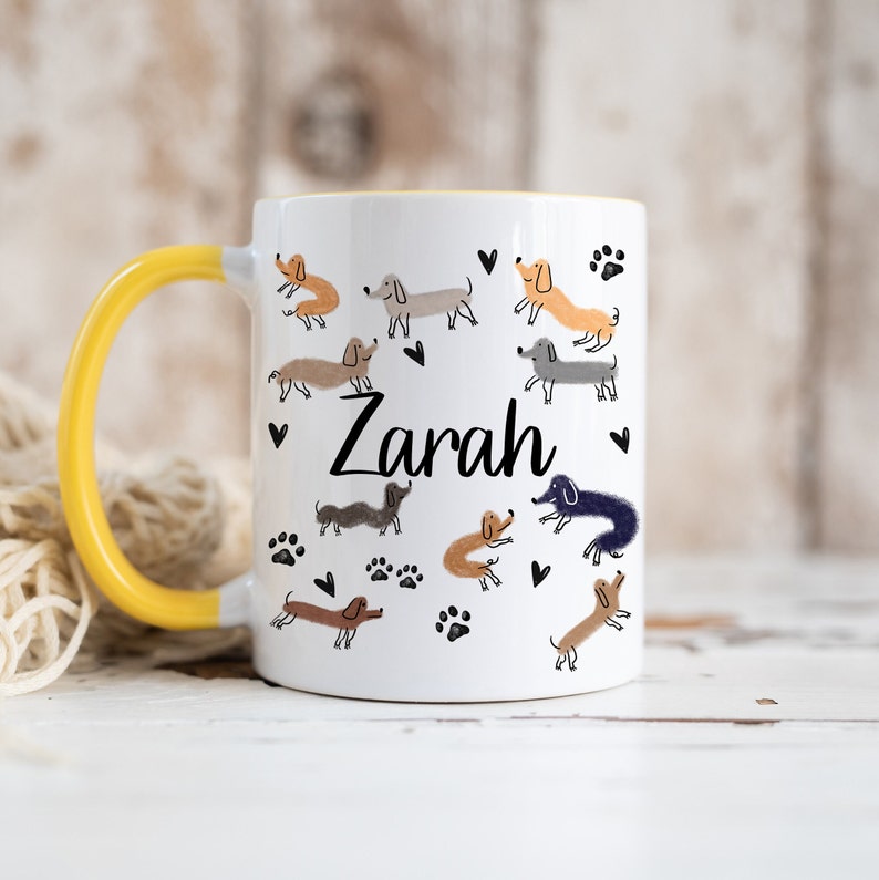 Pode incluir: Uma caneca de cer&acirc;mica branca com uma al&ccedil;a amarela brilhante, apresentando um padr&atilde;o de c&atilde;es dachshund de v&aacute;rias cores, pegadas pretas e pequenos cora&ccedil;&otilde;es. O nome Zarah est&aacute; impresso em preto no centro do design da caneca.