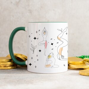 May include: White ceramic mug with a green handle and rim, featuring line art illustrations of yoga poses. The design includes stars and leaves. The mug is surrounded by gold coins and green beads.