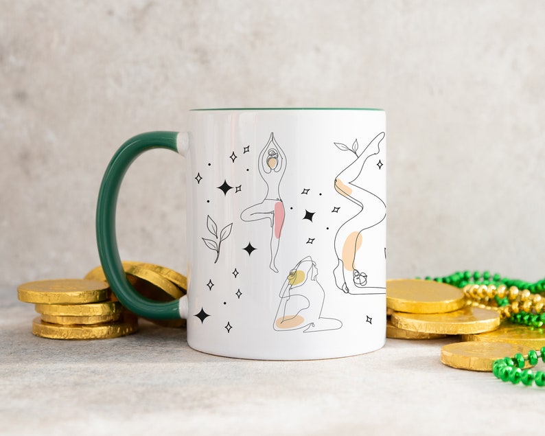 May include: White ceramic mug with a green handle and rim, featuring line art illustrations of yoga poses. The design includes stars and leaves. The mug is surrounded by gold coins and green beads.