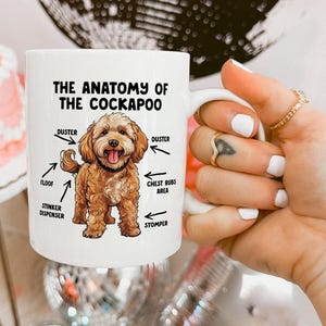 Peut inclure: Une tasse en céramique blanche avec une illustration d'un chien Cockapoo brun et blanc. La tasse porte le texte "The Anatomy of the Cockapoo" avec des étiquettes pour "Duster", "Floof", "Stinker Dispenser", "Chest Rubs Area" et "Stomper".