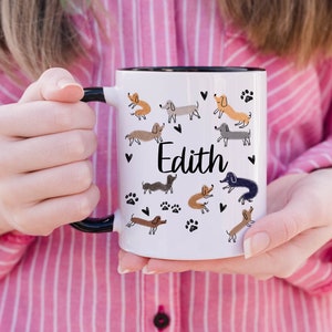Pode incluir: Uma caneca de cer&acirc;mica branca com asa e rebordo pretos, apresentando um padr&atilde;o repetitivo de c&atilde;es dachshund de v&aacute;rias cores, pegadas pretas e pequenos cora&ccedil;&otilde;es pretos. O nome Edith est&aacute; impresso em letra cursiva preta no centro.