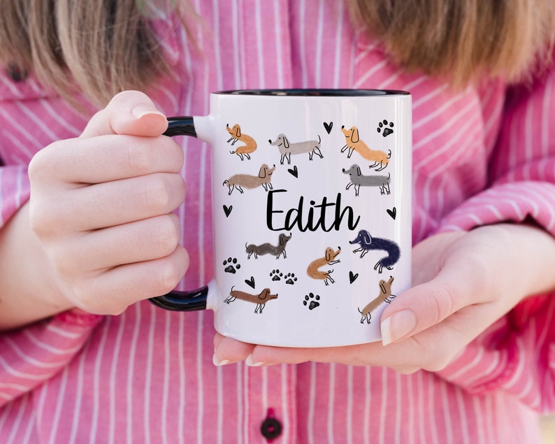 Pode incluir: Uma caneca de cer&acirc;mica branca com asa e rebordo pretos, apresentando um padr&atilde;o repetitivo de c&atilde;es dachshund de v&aacute;rias cores, pegadas pretas e pequenos cora&ccedil;&otilde;es pretos. O nome Edith est&aacute; impresso em letra cursiva preta no centro.