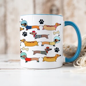 May include: White ceramic coffee mug with a blue rim and handle. The mug features a repeating pattern of dachshund dogs in various costumes, including a hot dog, a beret, and an astronaut helmet.