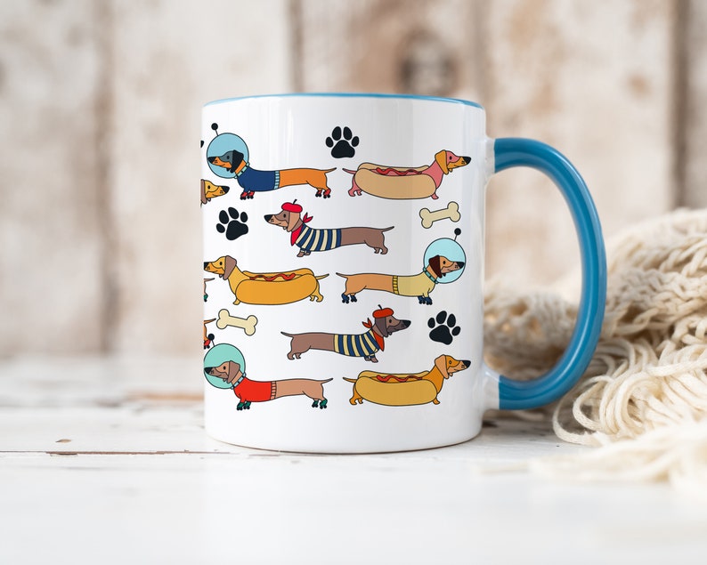 May include: White ceramic coffee mug with a blue rim and handle. The mug features a repeating pattern of dachshund dogs in various costumes, including a hot dog, a beret, and an astronaut helmet.