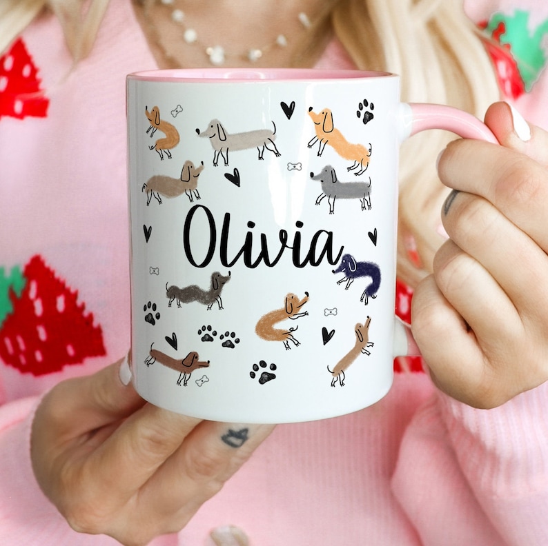 Pode incluir: Uma caneca de cer&acirc;mica branca com interior e al&ccedil;a rosa claro, apresentando um padr&atilde;o repetitivo de ilustra&ccedil;&otilde;es de c&atilde;es dachshund em tons de marrom, cinza e preto, pegadas, cora&ccedil;&otilde;es e ossos pretos, com o nome Olivia em letra cursiva preta.