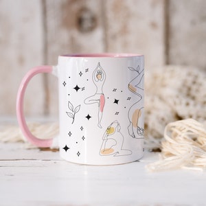 May include: White ceramic mug with a pink handle and interior. The mug features line art illustrations of yoga poses, with accents of pink, yellow, and black. The design includes stars and leaves.