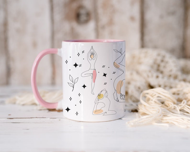 May include: White ceramic mug with a pink handle and interior. The mug features line art illustrations of yoga poses, with accents of pink, yellow, and black. The design includes stars and leaves.