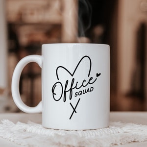 May include: A white ceramic mug with a black graphic print featuring the text Office SQUAD in a stylized script and sans-serif font, accompanied by a heart outline and a small cross symbol. The mug has a curved handle and a smooth finish.