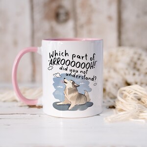May include: A white ceramic mug with a pink handle and interior rim. The mug features a graphic of a howling dog and the text: Which part of ARROOOOOOH! did you not understand? The dog is light brown and white, positioned against a blue sky.