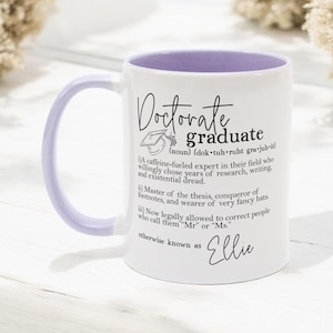 Könnte beinhalten: Weißer Keramikbecher mit hellviolettem Griff und Innenraum. Der Becher trägt die Aufschrift "Doctorate graduate" mit einer Definition und dem Namen "Ellie". Ein tolles Geschenk für einen Absolventen.
