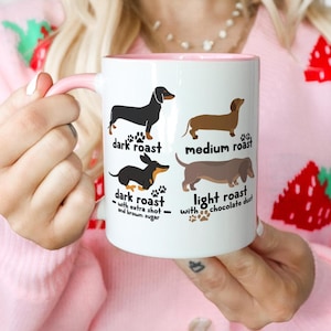 May include: White ceramic coffee mug with a pink interior and handle. The mug features illustrations of dachshunds labeled "dark roast," "medium roast," and "light roast." Additional text reads "with extra shot and brown sugar" and "with chocolate dust."