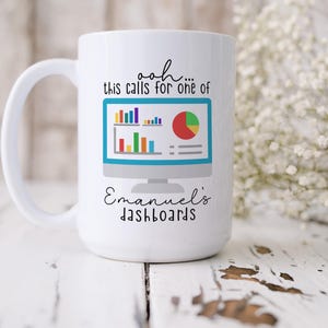 May include: A white ceramic mug featuring a graphic of a computer monitor displaying colorful bar and pie charts. The text above the monitor reads "ooh... this calls for one of" and the text below reads "Emanuel's dashboards" in black script and print.