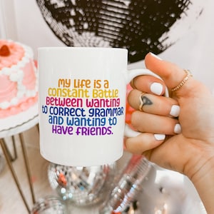 Peut inclure: Une tasse en céramique blanche avec un motif coloré qui dit "Ma vie est une bataille constante entre le désir de corriger la grammaire et le désir d'avoir des amis."