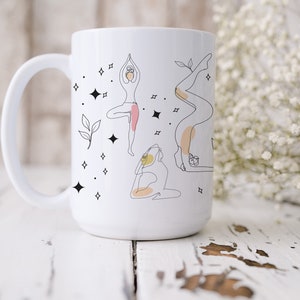 May include: White ceramic mug with line art illustrations of yoga poses in black, with pink and peach accents. The design includes stars and leaves. The mug has a large handle and is set against a white wooden surface.